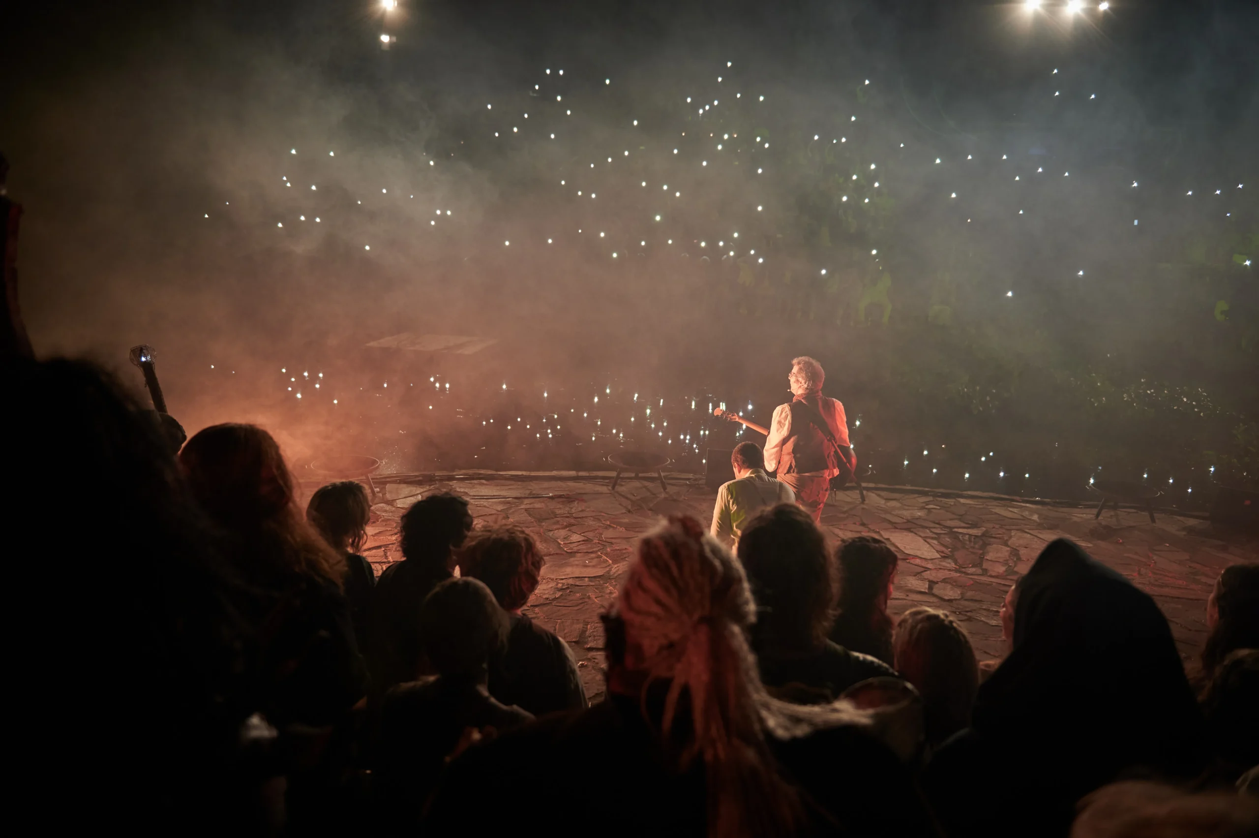 Bilbo Beutlin steht auf der Bühne, mit Blick in Richtung des Publikums im Hintergrund. Das Publikum hat die Handy-Taschenlampen eingeschaltet und man sieht ein Lichtermeer.
