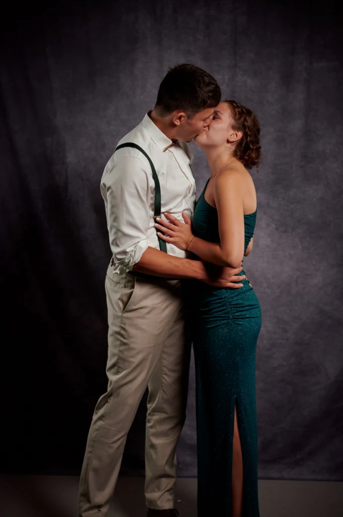 Ein junger Mann hält seine Freundin im Arm und sie küssen sich. Beide stehen in einem Fotostudio vor grau meliertem Hintergrund.